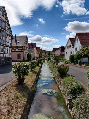 Blick entlang eines offenen Wasserkanals in der Mitte zwischen zwei Häuserzeilen. Au beiden Seiten des Wasser geht eine Straße entlang, die von Haus zu Haus führt. Links stehen einige Fachwerkhäuser, rechts schlichte Wohnhäuser aus der Nachkriegszeit.