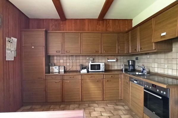 Eine große L-förmige Holz-Küchenzeile mit verschieden Elektrogeräten. Seiten- und Rückwand der Küche sind holzvertäfelt. Zwei Balken verlaufen durch die helle Decke.