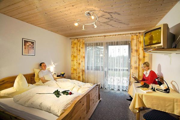 Schlafzimmer mit grauem Teppichboden, Holzdecke sowie Balkontür und - fenster mit gelb gemusterten Vorhängen. Im Doppelbett im Landhausstil liegt ein Mann, der seine am Tisch sitzende Partnerin anlächelt. Auf dem Tisch eine Rotweinflasche und zwei Gläser, darüber ein Fernseher.
