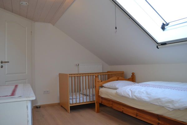 Unter einer Mansarde mit Dachfenster steht ein Kieferholz-Einzelbett, dahinter ein Kindergitterbett; gegenüber eine weiße Kommode mit Tischdeckchen.