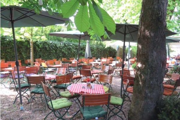 Laubbäume, Sonnenschirme und eine Hecke spenden Schatten im Biergarten. Auf der Kiesfläche stehen runde und eckige Gartentische, gedeckt mit rot-weiß karierten Tischdecken, und Stühle mit Sitzauflagen in verschiedenen Grüntönen.