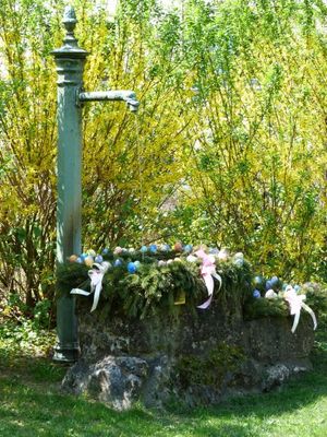 Unter dem Gußeisernen Wasserauslauf steht ein steinerner Brunnen auf dessen Rand eine grüne Girlande mit Ostereiern angebracht ist.