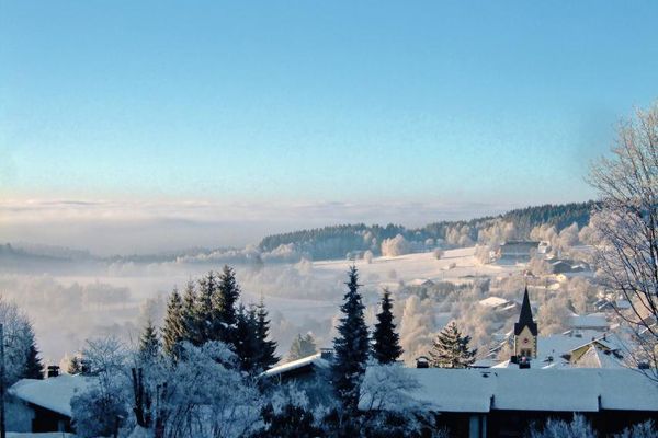 Schöner Blick über Sankt Englmar mit Kirchturm in eine weite, sonnige Winterlandschaft, die teils leicht im Dunst liegt.