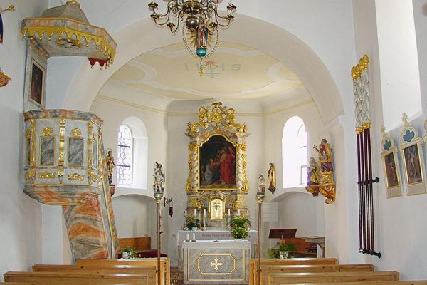 Heller Kircheninnenraum mit beidseitigen Bankreihen, Kanzel, Altar und einigen Heiligenfiguren.