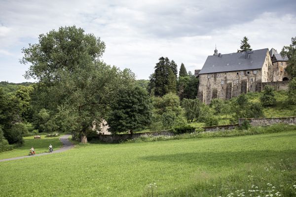 Zwei Radfahrer fahren vor Schloss Eisenbach, das auf einem Bergsporn thront, auf einem asphaltierten Radweg. Die Umgebung ist herrlich grün.