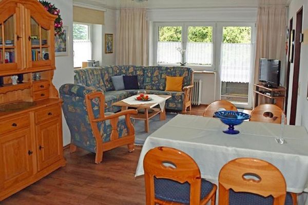 Wohnzimmer im Landhausstil: Polstereckbank, Holztisch, Sessel, Holzbüffet, Esstisch mit vier Stühlen. Ein TV-Gerät steht auf einem Eckregal neben der Terrassentür.