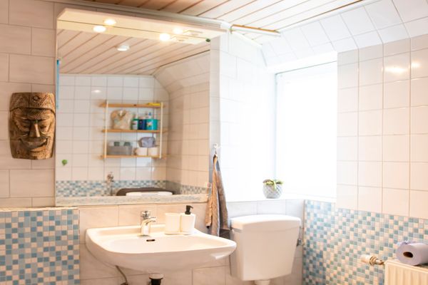 Ein Badezimmer mit einem weißen Waschbecken, einem großen beleuchteten Spiegel und einer weißen Toilette. Die Wände sind oben mit weißen Kacheln und unten mit blau-weißen Mosaikfliesen bedeckt. An der linken Wand hängt eine geschnitzte Holzmaske, während rechts helles Tageslicht durch ein Fenster fällt.