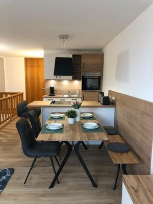 Die Wand hinter dem Essbereich ist halbhoch mit Holz verkleidet. Davor stehen eine Bank, ein Tisch und zwei graue Stühle. Der Tisch ist für vier Personen gedeckt.Direkt anschließend befindet sich eine Esstheke mit eingebautem Ceranfeld, beleuchtet von einer dunklen Deckenlampe. Vor der Theke stehen zwei weitere graue Stühle. Dahinter ist eine moderne Küche in Weiß und Holz mit einem Backrohr in Sichthöhe.