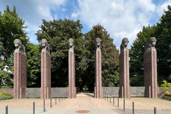 Sechs hohe Backsteinsäulen mit Löwenköpfen als Abschluss stehen in einer Parkanlage vor einem Weg, im Hintergrund Bäume und blauer Himmel mit Wolken