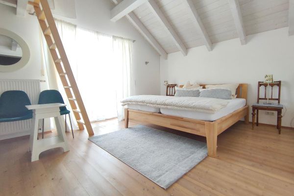 Vor einer Fensterfront und unter einem offenen, hellen Dachstuhl steht ein Doppelbett mit Holzrahmen zwischen zwei Stühlen mit Lampen. Vor dem Bett liegt ein heller Teppich. An der Wand stehen zwei blaue Stühle unter einem ovalen Spiegel. Eine Holztreppe führt hinauf zur Galerie.