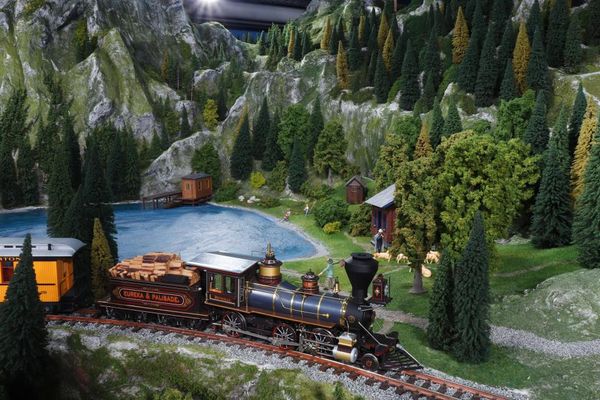 In kanadischer Berglandschaft zieht eine historische Lok, vorbei an einem See, einige Wagons, die zum Teil mit Gütern beladen sind.