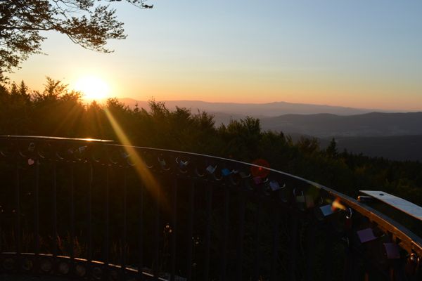 Ein fantastischer Sonnenaufgang über den Wäldern und Hügeln in der Ferne, im Vordergrund das Geländer des Hirschenstein-Aussichtsturms.