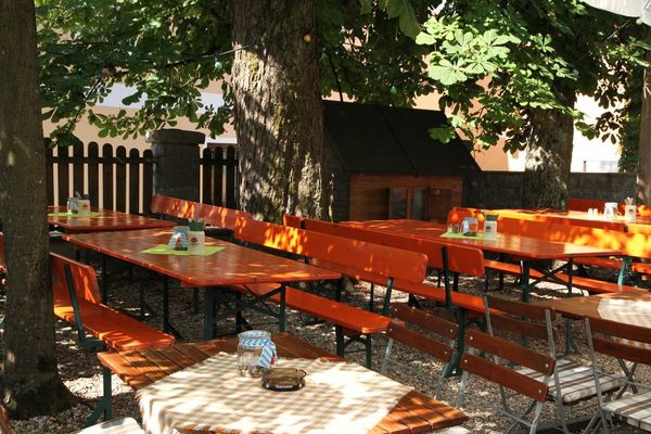 Ein Biergarten unter schattigen Bäumen. Auf den Biertischen stehen Steinkrüge mit Servietten und Besteck.