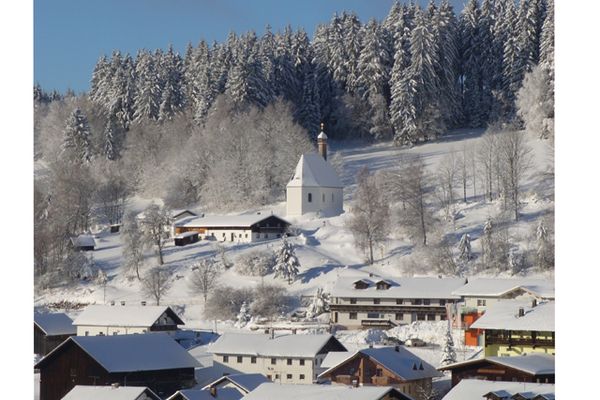 Winterliche Idylle: Im Vordergrund stehen einige schneebedeckte Häuser von Sankt Englmar, dahinter erhebt sich die Leonhardikapelle vor einem verschneiten Wald.Im Winterwald steht im
