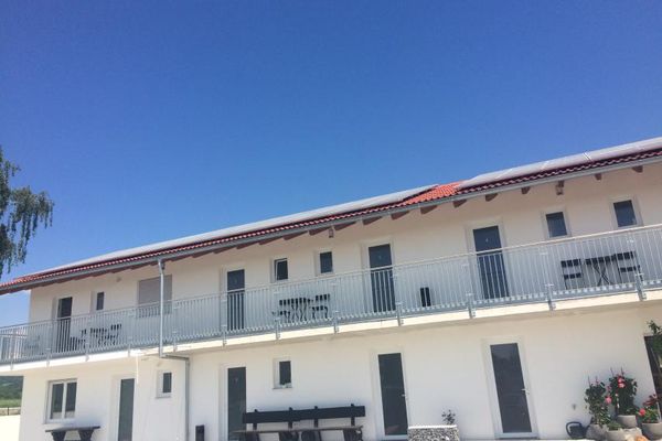 Ein langes, weißes Gebäude mit Edelstahlbalkonen, mehreren Balkon- und Terrassentüren sowie kleinen Fenstern. Auf den Terrassen stehen ein Tisch, eine lange Bank und drei runde Steintröge mit Blumen.