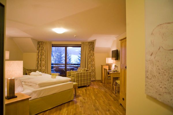Im Raum steht beiges Doppelbett, die Fenster sind bodentief mit beige gemusterten Vorhängen, Zugang zum Balkon