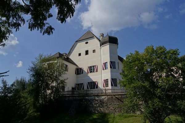 Oberhalb einer Steinmauer und hinter einem niederen Lattenholzzaun ragt das Schloss Haggn in den weiß blauen Himmel. Auf der Wiese vor der Anlage stehen einige Laubbäume.