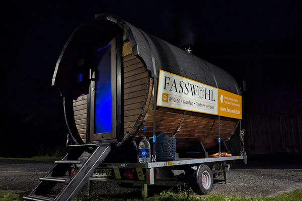Auf dem Bild ist der Anhänger mit Fass-Sauna bei Dunkelheit zu sehen.