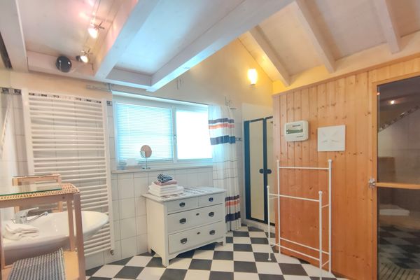 Ein Badezimmer mit schwarz-weißen Bodenfliesen unter einem offenen, hellen Dachstuhl. Es gibt ein Waschbecken, einen großen Handtuchheizkörper, eine weiße Kommode unter einem Doppelfenster, eine Dusche und eine Sauna.