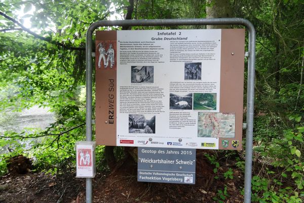 Eine Infotafel im Wald zum Erzweg Süd und zur Grube Deutschland. Unter der Tafel ist ein Schild mit dem Zusatz Geotop des Jahres 2015 und Weickartshainer Schweiz angebracht.