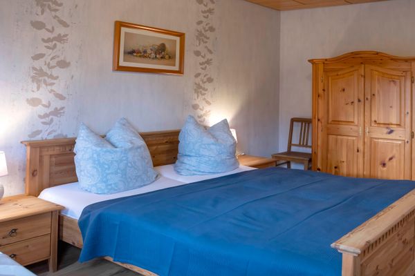 Blick in ein Schlafzimmer mit hellen Holzmöbeln. Im Vordergrund steht ein Doppelbett mit einer dunkelblauen Tagesdecke und hellblau gemusterten Kopfkissen. Links und rechts stehen passende Nachttische mit Lampen. Im Hintergrund rechts steht ein zweitüriger Kleiderschrank aus Holz, daneben ein Holzstuhl. An der Wand hängt ein gerahmtes Blumenbild.