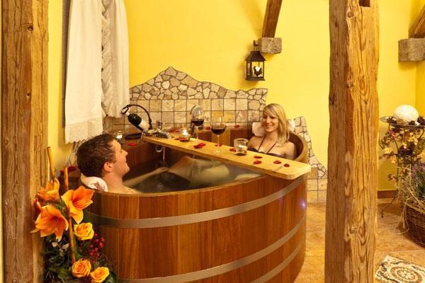 Baderaum mit alten Holzpfeilern und gelber Wand, die teils mit Mosaikfliesen verziert ist. Im ovalen Holzzuber badet ein Pärchen. Quer über dem Zuber liegt ein Brett, darauf befinden sich zwei Gläser mit Rotwein und roten Blütenblätter. Blumendeko schmückt den Raum.