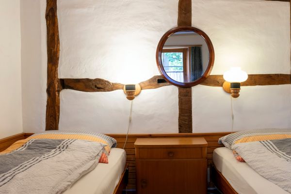 Zwei Einzelbetten mit Nachttisch dazwischen, Wand mit Holzbalken, Spiegel und zwei Lampen.