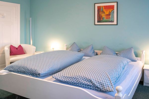 Weißes Doppelbett mit blauer Bettwäsche vor einer türkisfarbenen Wand. Links und rechts stehen weiße Nachttische mit eingeschalteten Lampen. Links im Hintergrund steht ein weißer Sessel mit rotem Kissen vor einer weißen Tür. Rechts oben hängt ein gerahmtes Bild.