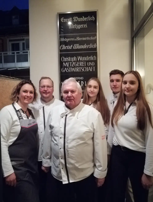 Auf dem Bild ist das Team der Gaststätte zu sehen. 6 Personen sind vor einer Tafel an der Wand stehend zu sehen. Sie tragen Arbeitskleidung (Koch, Bedienung).