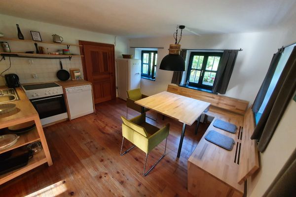 Wohnküche mit dunklen Sprossenfenstern, Holzeckbank, passendem Tisch und zwei grünen Stühlen. Spüle, Herd und Spülmaschine in L-Form. Darüber zwei Regale mit Deko und Küchenutensilien. Ein Gefrierschrank steht separat.