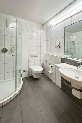 Das Bild zeigt ein modernes Badezimmer mit einer Dusche, einer wandmontierten Toilette und einem Waschbecken. Die Wände sind mit weißen Fliesen verkleidet und der Boden ist grau gefliest. Über dem Waschbecken befindet sich ein großer Spiegel.