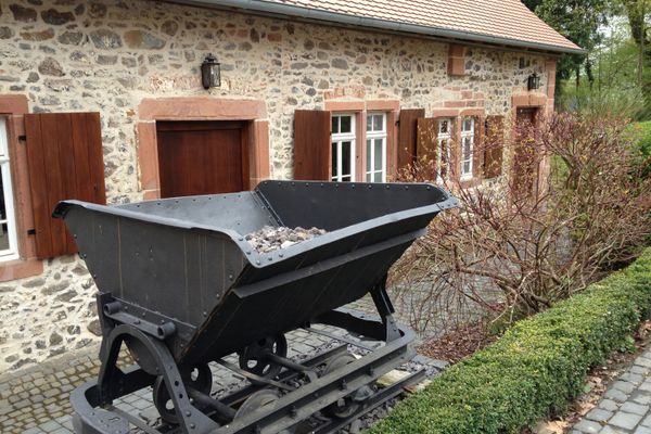 Eine schwarze, metallerne Lore mit Basaltgestein gefüllt steht vor einem Steinhaus auf einem kurzen Schienenstück.