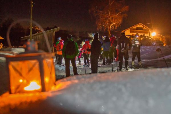 Eine Gruppe Schneeschuhwanderer steht auf einem verschneiten Weg, bereit zum Aufbruch. Links im Vordergrund steckt eine steckt im Schnee im Vordergrund. Hinter der Gruppe leuchtet an einem Haus das Hoflicht.