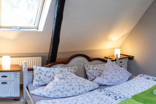 Ein helles Schlafzimmer mit weißer Dachschräge und einem dunklen Holzbalken im Raum. In der Mitte steht ein Doppelbett mit weiß-grau beblätterter Bettwäsche, an dessen Fußende eine leuchtend grüne Decke mit zwei aufgerollten Handtüchern liegt. Flankiert wird das Bett von zwei weißen Nachttischen mit Holzplatte, auf denen jeweils eine eingeschaltete eckige Lampe steht.