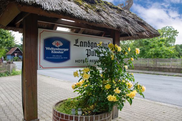 Überdachtes Schild mit Beschriftung und gelben Blumen in einem Pflanzgefäß am Straßenrand