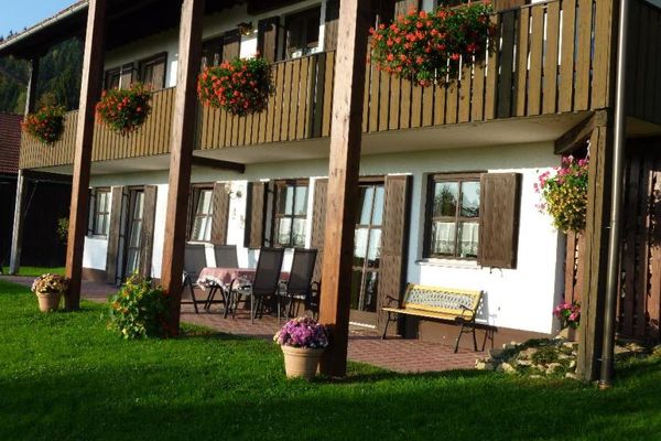 Teilansicht eines Hauses mit braunen Tür- und Fensterläden und Blumenschmuck auf Balkon und Liegewiese. Der Balkon wird durch mehrere Balken gestützt, darunter liegt die Terrasse auf der Gartenmöbeln und eine Bank stehen.