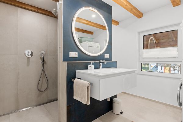 Badezimmer mit beigen Fliesen, an der Trennwand zwischen Dusche und Bad befindet sich ein Waschbecken mit Spiegel