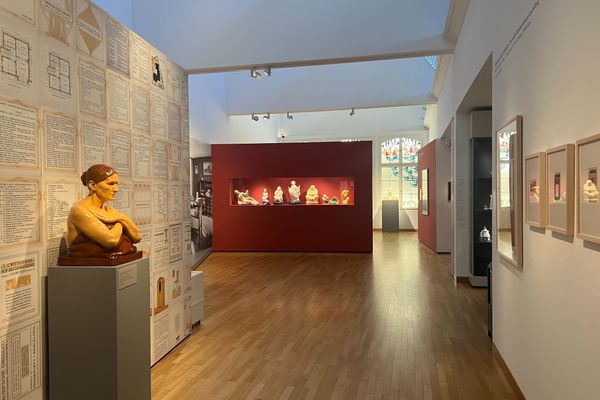 Museumsgang mit Holzfußboden, links Büste auf grauem Sockel vor Wand mit historischen Dokumenten, rechts gerahmte Bilder, im Hintergrund Vitrine mit mehreren kleinen Skulpturen.