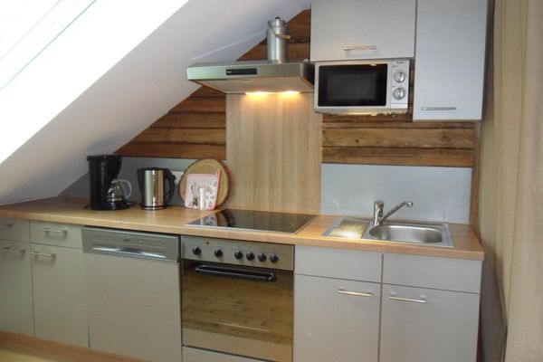 Unter einer Dachschräge mit Fenster steht eine helle Küchenzeile mit Mikrowelle; die Rückwand besteht teils aus rustikalem Holz.