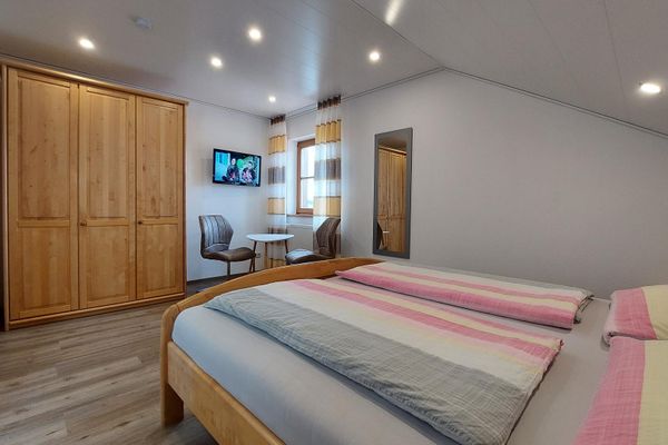 Ein Zimmer mit Doppelbett im Holzrahmen unter einer hellen Dachschräge. Gegenüber befinden sich ein Schrank, ein Flachbildfernseher und eine Sitzgruppe mit Tisch und zwei Stühlen vor einem Fenster mit bodenlangen, braun-beige gemusterten Vorhängen. An der Wand hängt ein Ganzkörperspiegel mit grauem Rahmen.