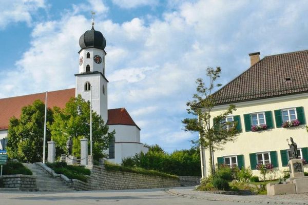 Steinstufen führen zu einer Kirche mit viereckigem Turm, Zwiebelkuppel und Steinmauer. Die Kirche ist umgeben von Sträuchern und Laubbäumen. Daneben ein großes Gebäude mit grünen Fensterläden und Blumenschmuck; davor ein Kriegerdenkmal.