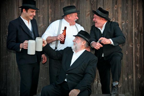 Vier Männer in schwarzem Anzug und weißem Hemd lachen sich herzlich an. Einer hat die Jacke ausgezogen. Sie prosten sich mit Steinbierkrügen und einer Bierflasche zu. Die Stimmung wirkt ausgelassen.