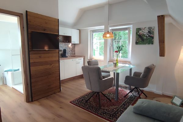 Wohnzimmer mit Couch, Flachbild-TV und Sitzecke. Im Hintergrund ist die Küche mit weißer Front zu sehen.