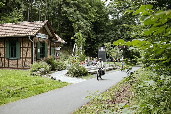 Radfahrer fahren auf dem geteerten Vulkanradweg an der Zentralstation in Lauterbach-Frischborn vorbei, die mitten in einem Waldstück liegt. Die Zentralstation ist der ehemalige Bahnhof und wird heute gastronomisch mit einem Biergarten genutzt.