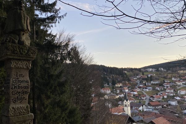 Im Vordergrund steht die Statue des Steinernen Engelmars idyllisch im Wald. Im Hintergrund liegt das Dorf Sankt Englmar.