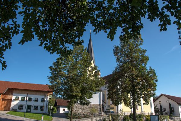 Die Dorfmitte von Haibach zeigt zwischen einigen Gebäuden die Kirche, die von hohen Bäumen etwas verdeckt wird und den Maibaum rechts im Vordergrund.