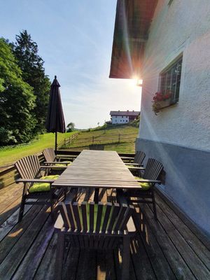 Vor dem Haus liegt eine Holzterrasse mit Tisch und Stühlen. Im Hintergrund Wiesen, ein weiteres Gebäude sowie ein stimmungsvoller Sonnenuntergang.