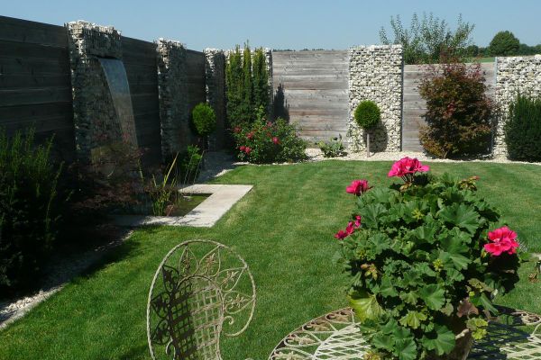 Eine auffällige Umzäunung aus hohen Gabionen, durchbrochen von verwitterten Holzbrettern und teils bewachsen mit hohen Pflanzen, umschließt einen Garten mit Rasenfläche. Im Vordergrund steht eine rot blühende Geranie auf einem runden Tisch.