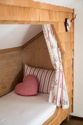 Mit heller Holz-Schindelwand verkleidetes Alkovenbett. Kissen und Vorhang im Landhausstil. An der Schindelwand hängt ein kleines Geweih.