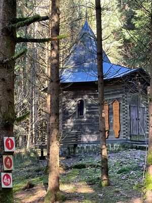 Mitten im Wald steht eine kleine Holzkapelle mit auffälligem Dach. Davor eine Holzbank, an der Wand zwei Totenbretter. Am einem Baum befinden sich drei Hinweisschilder.